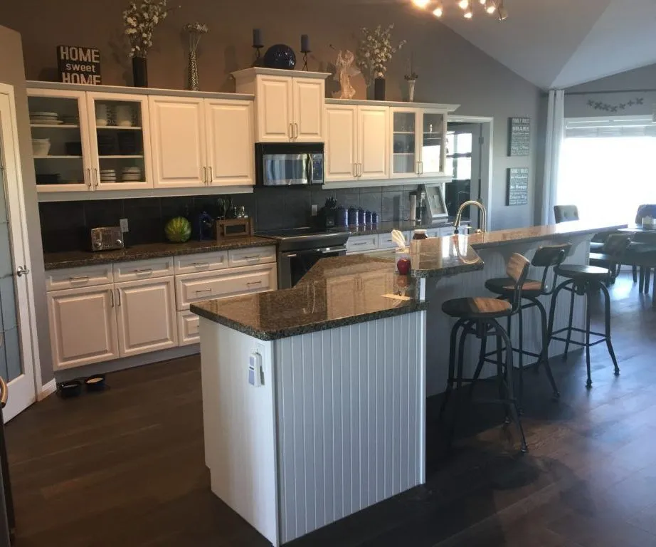 Modern kitchen with white cabinets, granite countertops, and a central island. Three bar stools line the breakfast bar, and decor items adorn the top.