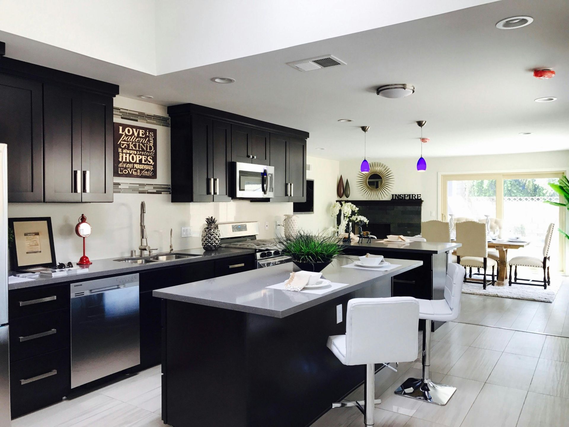 Modern kitchen and dining area with dark cabinets, stainless steel appliances, island with bar stools, purple pendant lights, and dining table